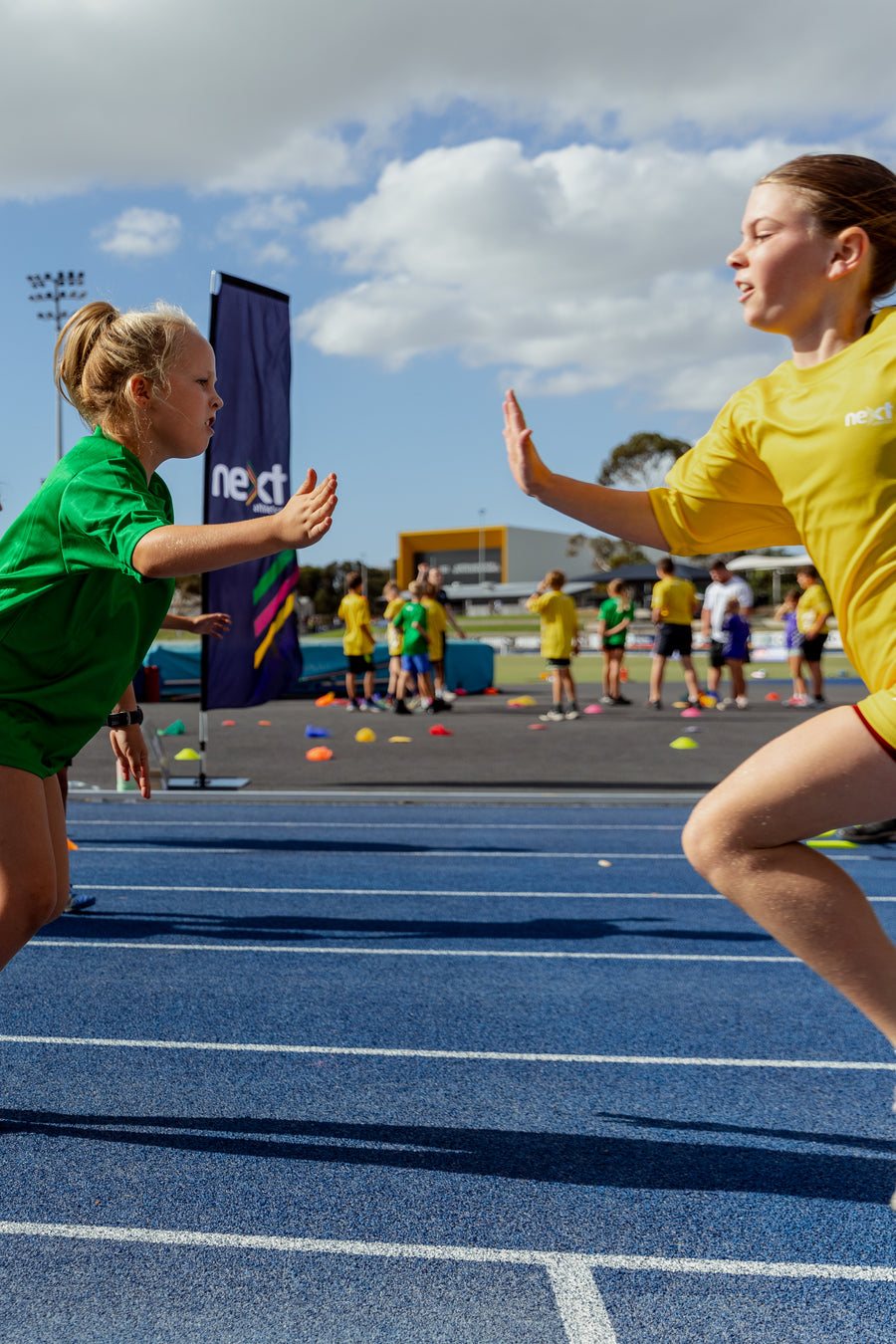 Two kids on a track and field event, one in green and one in yellow, with a blue track and cloudy sky in the background.