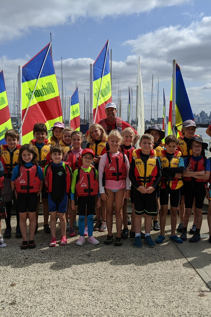 A group of children wearing life jackets pose together outdoors in front of colorful sailboats and flags on a sunny day at an ASC learn-to-sail program.