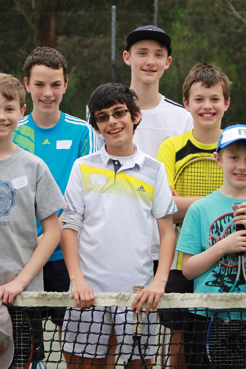 A group of seven young boys at an ASC tennis camp, smiling and posing together on a tennis court behind a net and holding tennis rackets.
